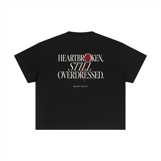 Back of the black Night Receipt boxy cotton tee with HEARTBROKEN, STILL OVERDRESSED in editorial serif capitals and a pressed oxblood rose blooming through the letterform