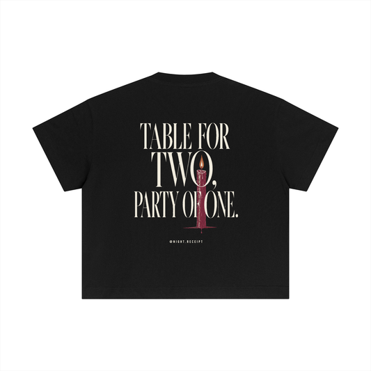 Back of the black Night Receipt boxy cotton tee with TABLE FOR TWO, PARTY OF ONE in editorial serif capitals and a single lit oxblood taper candle threading the O of TWO and the O of ONE