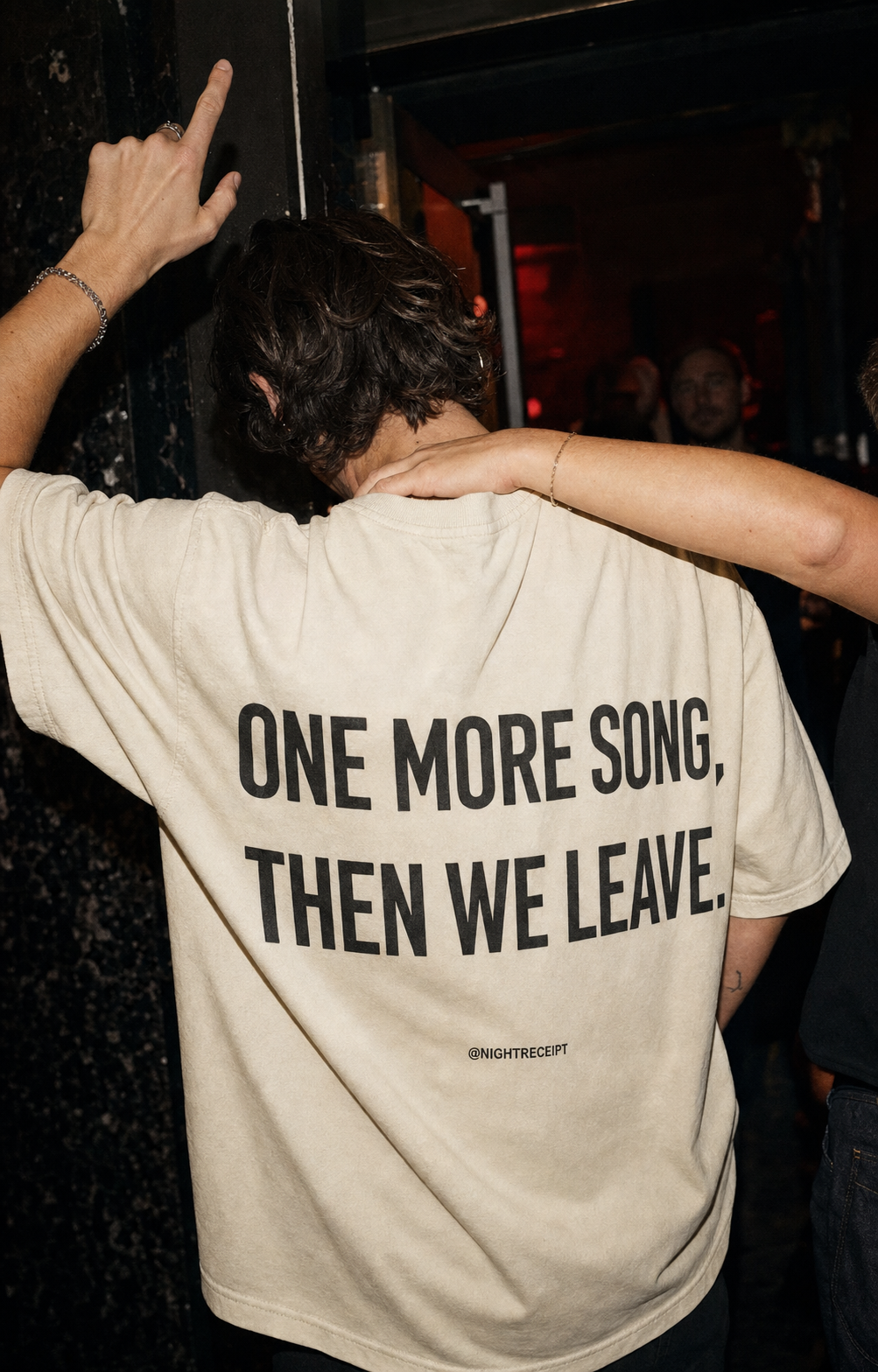 Person in a dark club wearing the cream Night Receipt oversized tee with the ONE MORE SONG, THEN WE LEAVE back print