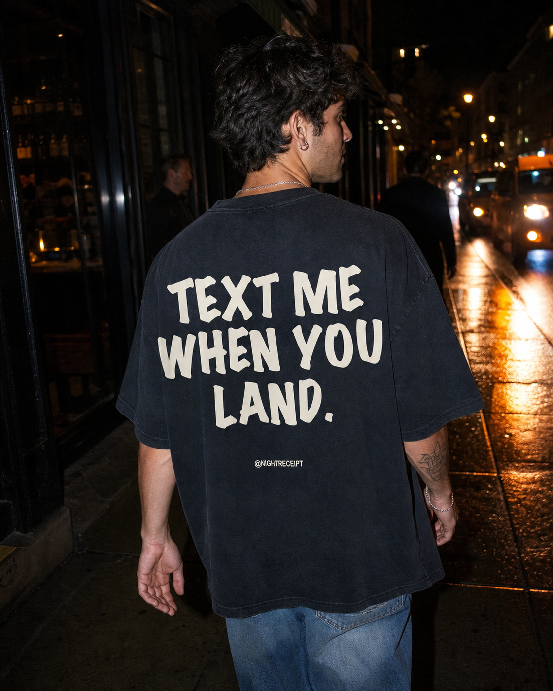 Man on a rain-slicked night street wearing the black Night Receipt oversized tee, cream TEXT ME WHEN YOU LAND back print